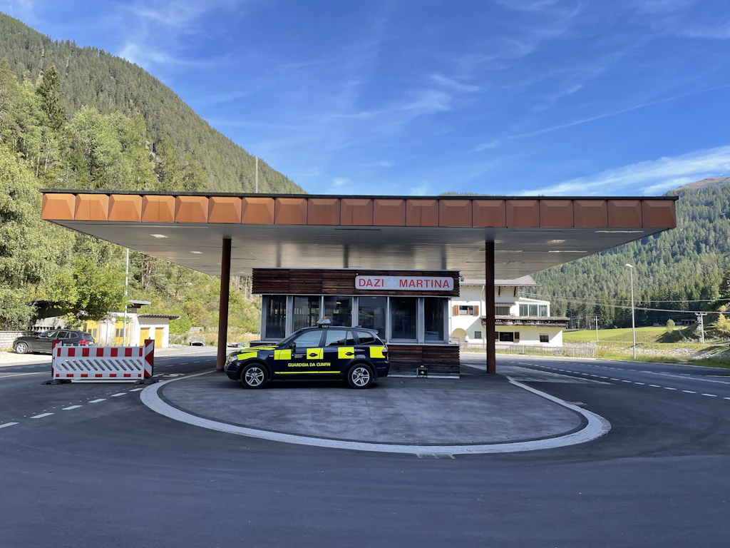 Un edificio doganale al valico di Martina con un veicolo di servizio della guardia di confine in primo piano. Sullo sfondo si vedono abitazioni, boschi e montagne.  